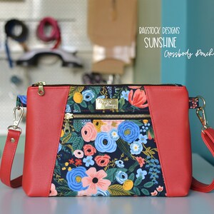 Op de afbeelding: Rode crossbody tas met een bloemenprint en gouden ritsen. De tas heeft de tekst "Bagstock Designs Sunshine Crossbody Pouch" op de voorkant.