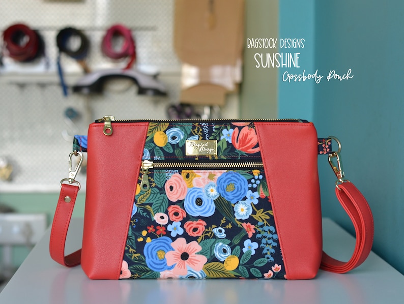 Op de afbeelding: Rode crossbody tas met een bloemenprint en gouden ritsen. De tas heeft de tekst "Bagstock Designs Sunshine Crossbody Pouch" op de voorkant.