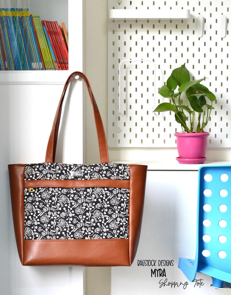 May include: A brown leather tote bag with a black and white floral print fabric lining. The bag has a zippered pocket on the front and a top zipper closure. The bag is labeled "Bagstock Designs Myra Shopping Tote".