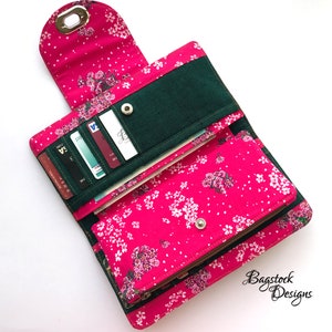 May include: A pink wallet with a floral print and a green fabric flap. The wallet is open and shows multiple card slots, a zippered compartment, and a billfold. The wallet has a snap closure and a metal ring.