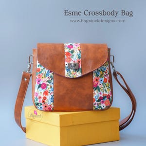 Op de afbeelding: Een bruine kunstleren schoudertas met een klep van bloemenstof en een zilveren sluiting. De tas ligt op een gele doos. De tekst "Esmé Crossbody Bag" en "www.bagstockdesigns.com" is zichtbaar boven de tas.