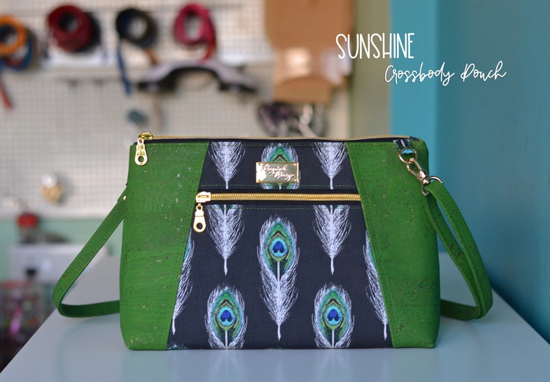 Op de afbeelding: Een groene en zwarte crossbody tas met een ritssluiting en een pauwenverenprint. De tas heeft een gouden rits en een gouden metalen label met de tekst "Design by Daisy". De tekst "SUNSHINE Crossbody Pouch" is in wit op een blauwe achtergrond geschreven.