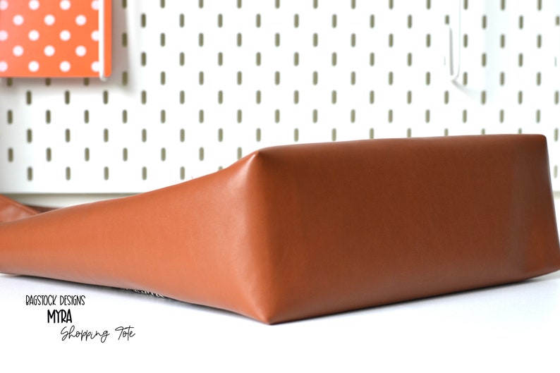 May include: A brown leather tote bag with the text "BAGSTOCK DESIGNS MYRA Shopping Tote" printed on the front.