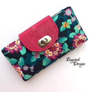 May include: A pink and teal floral print wallet with a cork and pink flap closure and a gold metal clasp. The wallet has a zipper on the top.