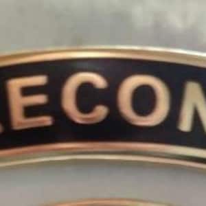 May include: A gold-colored metal patch with a black background and the word "RECON" in gold letters.