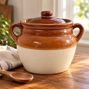 May include: A two-toned ceramic bean pot with a glossy brown glazed upper section and lid, and a matte cream-colored lower base. The pot features two side handles and a rounded body shape, designed for cooking or serving food in a kitchen setting.