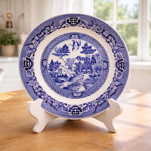 May include: A blue and white decorative plate with a Willow pattern design, displayed on a white stand. The plate features a detailed landscape scene and floral border. The plate is on a wooden table.