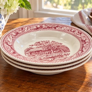 May include: A stack of three cream-colored ceramic soup bowls featuring a deep red transferware pattern. The rim displays an intricate border of oak leaves and acorns, while the center depicts a detailed rural landscape scene with a building.