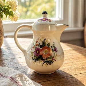 May include: Cream-colored ceramic pitcher with a lid, adorned with hand-painted floral designs in shades of red, purple, and green. The pitcher has a curved handle and a spout. It sits on a wooden table, next to a vase of flowers.
