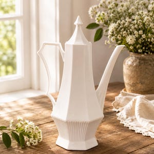 May include: A tall white ceramic coffee pot featuring a faceted hexagonal body, a pointed lid, a slender curved handle, and a long spout. The base of the vessel is decorated with vertical fluted ridges, providing a textured contrast to the smooth upper section.