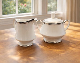 May include: A white porcelain creamer and a lidded sugar bowl with two handles. Both pieces feature a smooth, glossy finish and are accented with a thin, reflective silver metallic trim along the rims, bases, and the sugar bowl lid handle.