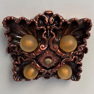 Puede incluir: Luminaria ornamentada de color cobre con cuatro bombillas globulares de vidrio esmerilado. La luminaria tiene un diseño esculpido detallado con un patrón floral o de hojas. Las bombillas están espaciadas uniformemente dentro del marco decorativo.