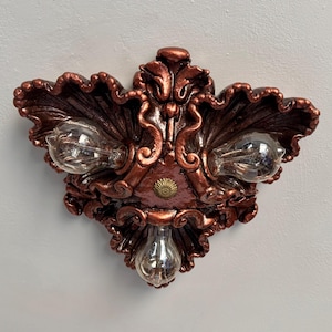 Puede incluir: Luminaria ornamentada de color cobre con tres bombillas expuestas. La luminaria tiene un diseño decorativo en forma de hoja con detalles intrincados y un pequeño acento circular en el centro.