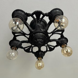 May include: Black, ornate ceiling light fitting with five exposed light bulbs. The fixture has a decorative, symmetrical design with curved arms and bulb holders. The light bulbs are round and have a warm, amber glow.