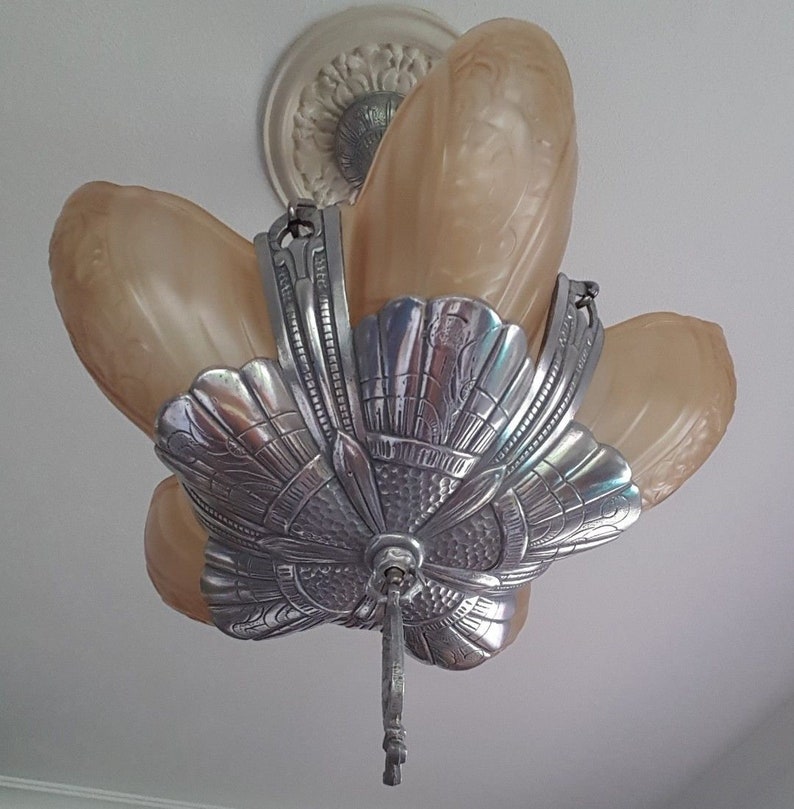 May include: A silver and glass ceiling light fixture with a decorative floral design. The fixture has five glass shades in a light brown color.