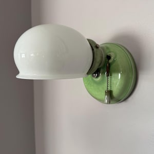 May include: A vintage-style wall sconce with a white, globe-shaped glass shade. The fixture has a round, green ceramic base and a pull chain. The sconce is mounted on a light-colored wall, offering a classic lighting solution.