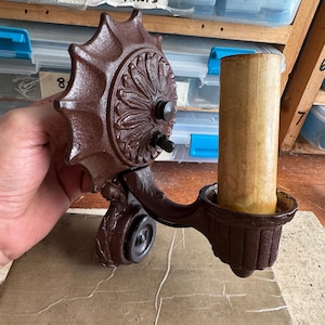 Puede incluir: Un candelabro marrón y ornamentado con un diseño decorativo de sol. El candelabro tiene un brazo curvo y una pequeña copa para sostener una vela. La vela es cilíndrica y de color beige. El artículo es de metal.