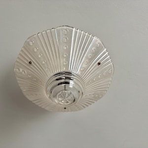 May include: A vintage ceiling light fixture with a clear, fan-shaped glass shade. The shade has a ribbed design with small, round accents and a central, metallic fixture. The light is mounted on a white ceiling.