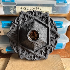 May include: A dark gray, ornate ceiling light fixture with a central light socket. The fixture has a detailed, symmetrical design with floral and geometric patterns. The fixture is sitting on a wooden surface.