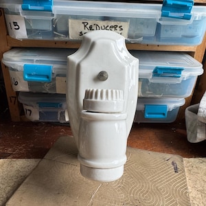 May include: A white ceramic object with a cylindrical base and a ridged top, possibly a bottle stopper or decorative piece. The object is set against a backdrop of plastic storage containers with blue clasps and labels.