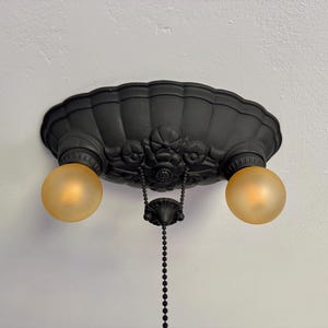 May include: Black, ornate ceiling light fixture with two round, amber-colored light bulbs. The fixture has a decorative, shell-like design and a pull chain. The light is mounted on a white ceiling.