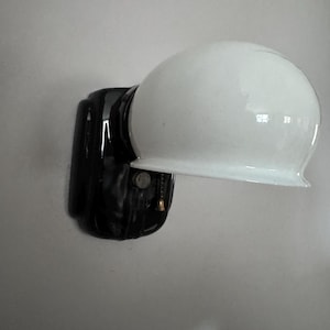 May include: A wall-mounted light fixture featuring a glossy black rectangular base and a smooth, rounded white glass shade. The fixture includes a small metallic pull chain with a bell-shaped end for operation, extending from the black base.