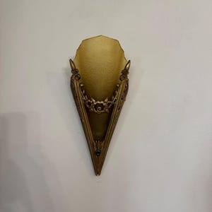May include: An antique wall sconce with a gold-toned metal frame and a frosted glass shade. The sconce has a triangular shape with ornate floral detailing and decorative accents. The sconce is mounted on a white wall.