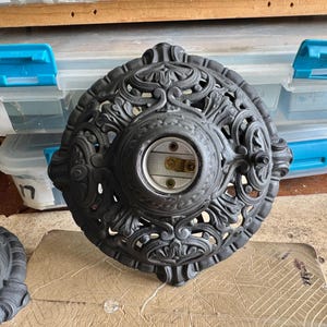 May include: Ornate, dark gray cast iron light fixture base with a central socket. The base features intricate floral and scrollwork designs. The fixture is likely vintage or antique, and is shown on a wooden surface.