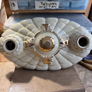 May include: Cream-colored, vintage-style light fixture with a scalloped design. Features two light sockets and a central decorative element with gold accents. The fixture is oval-shaped and appears to be made of ceramic or porcelain.