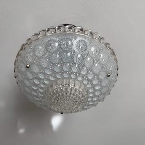 Pode incluir: Uma luminária de teto de vidro transparente com um design texturizado em forma de bolhas. A luminária redonda tem um padrão decorativo na parte inferior e é montada em uma base prateada.