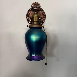 Puede incluir: Una lámpara de pared vintage con una pantalla de cristal azul y morado iridiscente. La lámpara tiene un accesorio de latón y una placa trasera decorativa de color cobre. Una cadena cuelga de la lámpara. La lámpara está montada en una pared blanca.