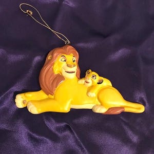 May include: A yellow and brown ceramic ornament of Simba and Mufasa from the Disney movie The Lion King. The ornament is shaped like the two lions lying down together.
