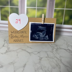 May include: A wooden sign with a white heart that says "24" on it. The sign says "Weeks Until Baby Morin Arrives". There is a black and white ultrasound image attached to the sign with a clothespin.