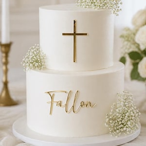 May include: A two-tiered white cake with gold cross and name accents. The top tier features a gold cross, while the bottom tier displays the name "Fallon" in gold script. The cake is decorated with small white flowers.