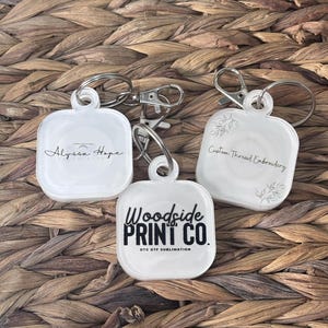 May include: Three white square keychains with different designs. The first keychain says "Alyssa Hope Photography". The second keychain says "Custom Thread Embroidery". The third keychain says "Woodside Print Co. DTG DTF Sublimation".