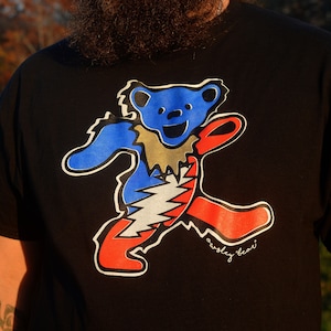 May include: Black t-shirt featuring a colorful dancing bear graphic. The bear is blue, red, silver, and gold with a white outline. The text "owsley bear" is written in cursive below the bear.