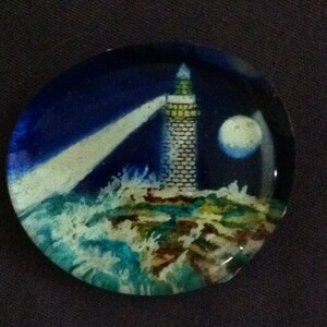 May include: A glass paperweight with a painted image of a lighthouse on a rocky shore with waves crashing against the rocks. The lighthouse is white with a black roof and a beam of light shining from the top. The sky is blue with a white moon.
