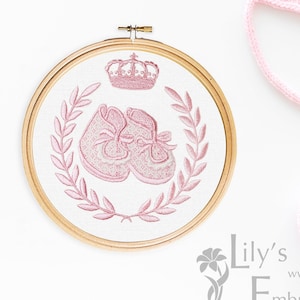 Può includere: Scarpine da bebè rosa e una corona ricamata su un tessuto bianco in un telaio da ricamo in legno. Il ricamo è circondato da una ghirlanda di foglie. Sono anche raffigurati un berretto e delle scarpine da bebè di lana rosa.