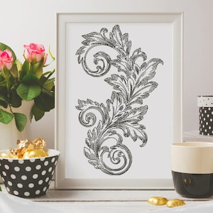May include: A black and white framed print of a decorative floral design with a white background. The print is sitting on a table with a white surface. There are pink roses in a white vase and a black and white polka dot bowl with gold chocolate candies on the table.