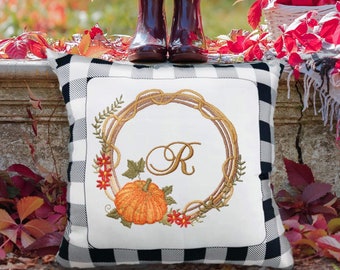 May include: A white and black checked cushion with an embroidered wreath featuring a pumpkin and the letter "R". The cushion is sitting on a stone surface with red leaves scattered around it.