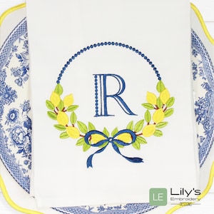 May include: Embroidered kitchen towel featuring a lemon wreath and a blue "R" monogram in the centre. The wreath is made of green leaves and yellow lemons. A blue bow is tied at the bottom of the wreath. The towel is white with a blue and white border.