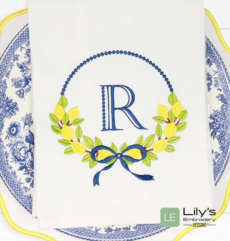 May include: Embroidered kitchen towel featuring a lemon wreath and a blue "R" monogram in the centre. The wreath is made of green leaves and yellow lemons. A blue bow is tied at the bottom of the wreath. The towel is white with a blue and white border.