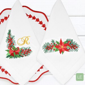 K&ouml;nnte beinhalten: Zwei wei&szlig;e bestickte Servietten mit Weihnachtsmotiv. Eine Serviette zeigt eine Weihnachtsstern-Blume und Kiefern&auml;ste, w&auml;hrend die andere einen Kranz aus Kiefern&auml;sten mit einer roten Schleife und dem Buchstaben "R" in Gold hat. Die Servietten liegen auf einer wei&szlig;en Holzoberfl&auml;che.
