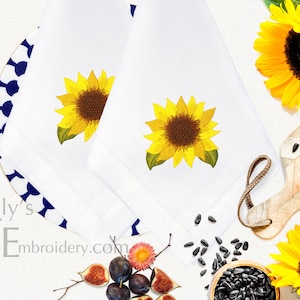 Pode incluir: Duas toalhas de cozinha brancas com girassóis bordados. As toalhas têm uma borda estampada em azul e branco. As toalhas são exibidas em uma superfície branca com sementes de girassol, uma tigela de sementes de girassol, um copo de óleo de girassol e um girassol.