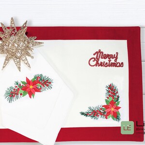 K&ouml;nnte beinhalten: Zwei wei&szlig;e bestickte Platzsets mit rotem Rand. Ein Platzset zeigt ein Design mit Weihnachtsstern und Tannenzweig, w&auml;hrend das andere den Text "Frohe Weihnachten" tr&auml;gt.