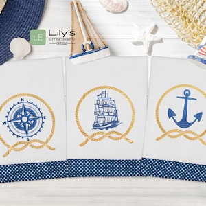 Könnte beinhalten: Drei weiße Geschirrtücher mit blauen nautischen Stickereien. Die Designs beinhalten einen Kompass, ein Schiff und einen Anker. Jedes Design ist von einem goldenen Seil umgeben.