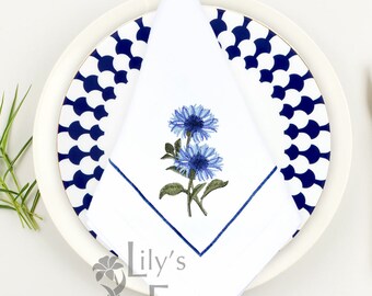 May include: White linen napkin with blue embroidered flowers and a blue border. The napkin is folded and placed on a white plate with a blue and white patterned border. The text "Lily's Embroidery" is visible in the bottom right corner.