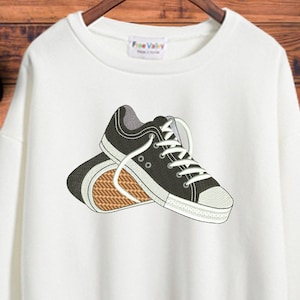 May include: White sweatshirt with an embroidered design of two black sneakers with white laces.