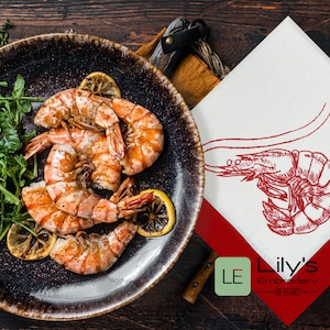 Puede incluir: Un plato de camarones cocidos con rúcula y gajos de limón sobre una superficie de madera marrón oscuro. Una servilleta blanca con ribete rojo presenta un diseño de camarón bordado en rojo y el texto "Lily's Embroidery Designs".