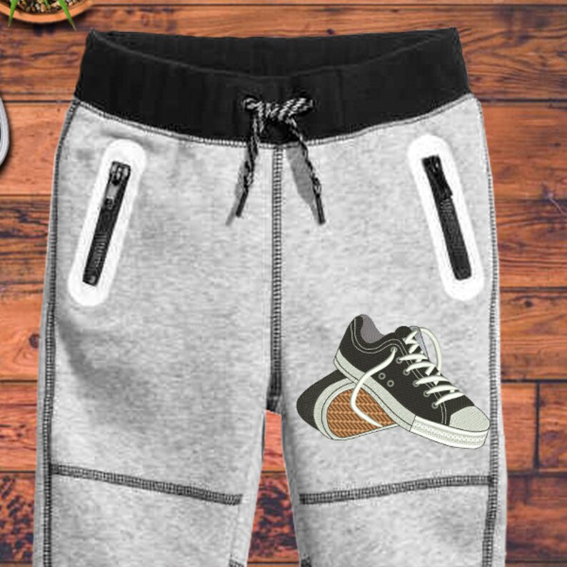 Puede incluir: Pantalones de ch&aacute;ndal grises con cintura negra y bolsillos con cremallera negros. Los pantalones de ch&aacute;ndal tienen un gr&aacute;fico de un par de zapatillas negras en la pierna derecha.